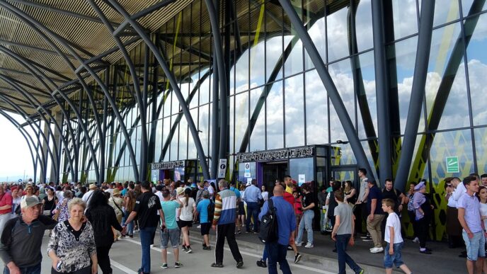 Pasageri în fața terminalului Aeroportului Internațional Brașov-Ghimbav, sub structura metalică ramificată a fațadei