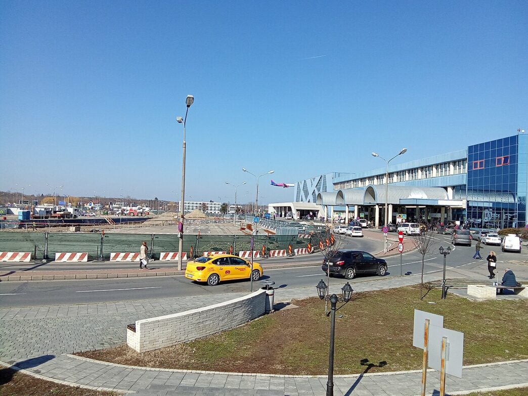 Clădirea terminalului Aeroportului Internațional Henri Coandă, văzută dinspre șoseaua de acces, cu un taxi galben și mai multe mașini pe carosabil