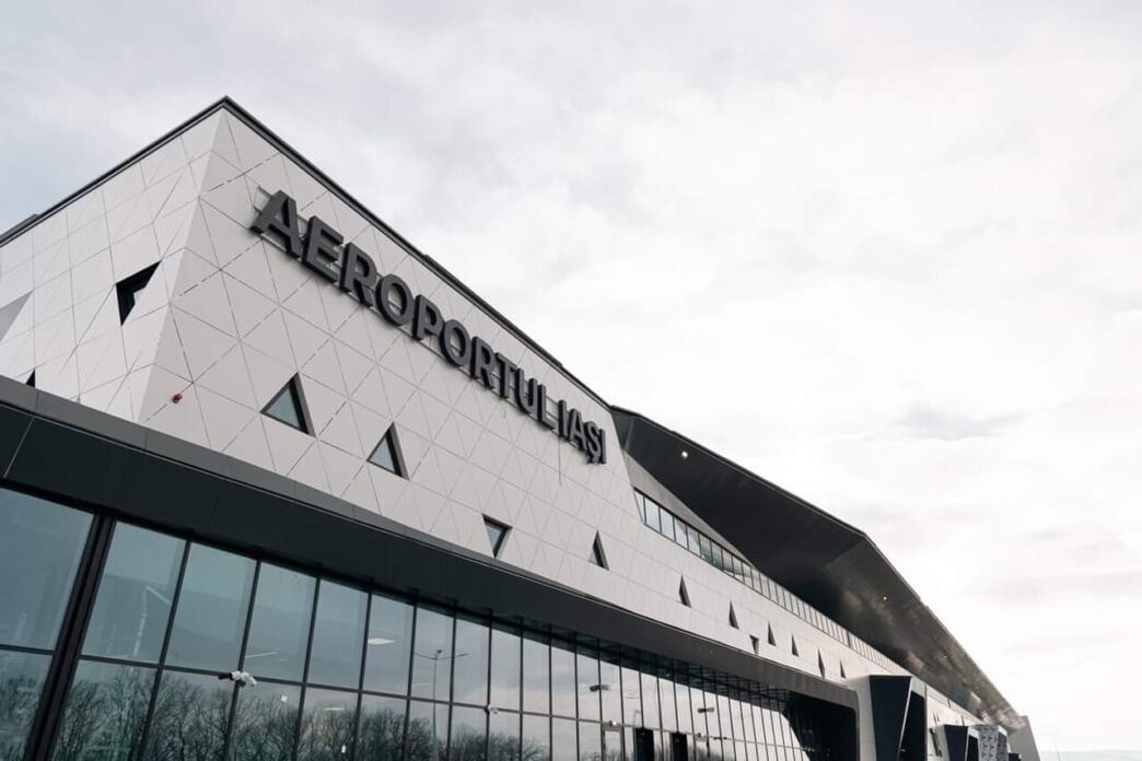 Fațada exterioară a unui terminal de aeroport, cu placare albă geometrică și litere mari montate pe perete, pe un cer acoperit