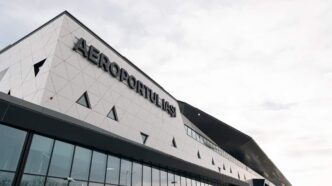 Fațada exterioară a unui terminal de aeroport, cu placare albă geometrică și litere mari montate pe perete, pe un cer acoperit