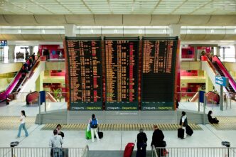 Panouri mari de afișaj pentru plecări într-un terminal de aeroport, cu pasageri și bagaje în prim-plan, în lumină de zi.
