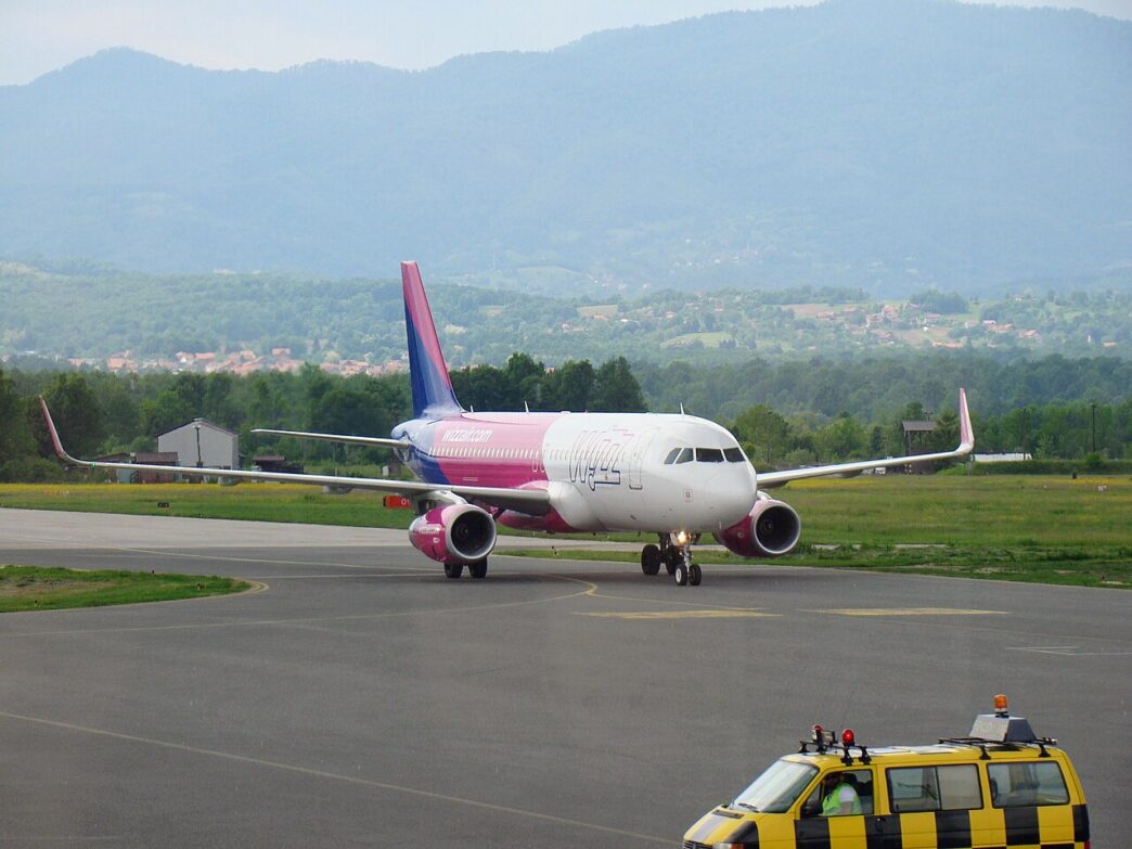 Aeronavă Airbus A320 în culorile Wizz Air, în rulare pe pista unui aeroport, cu dealuri împădurite în fundal